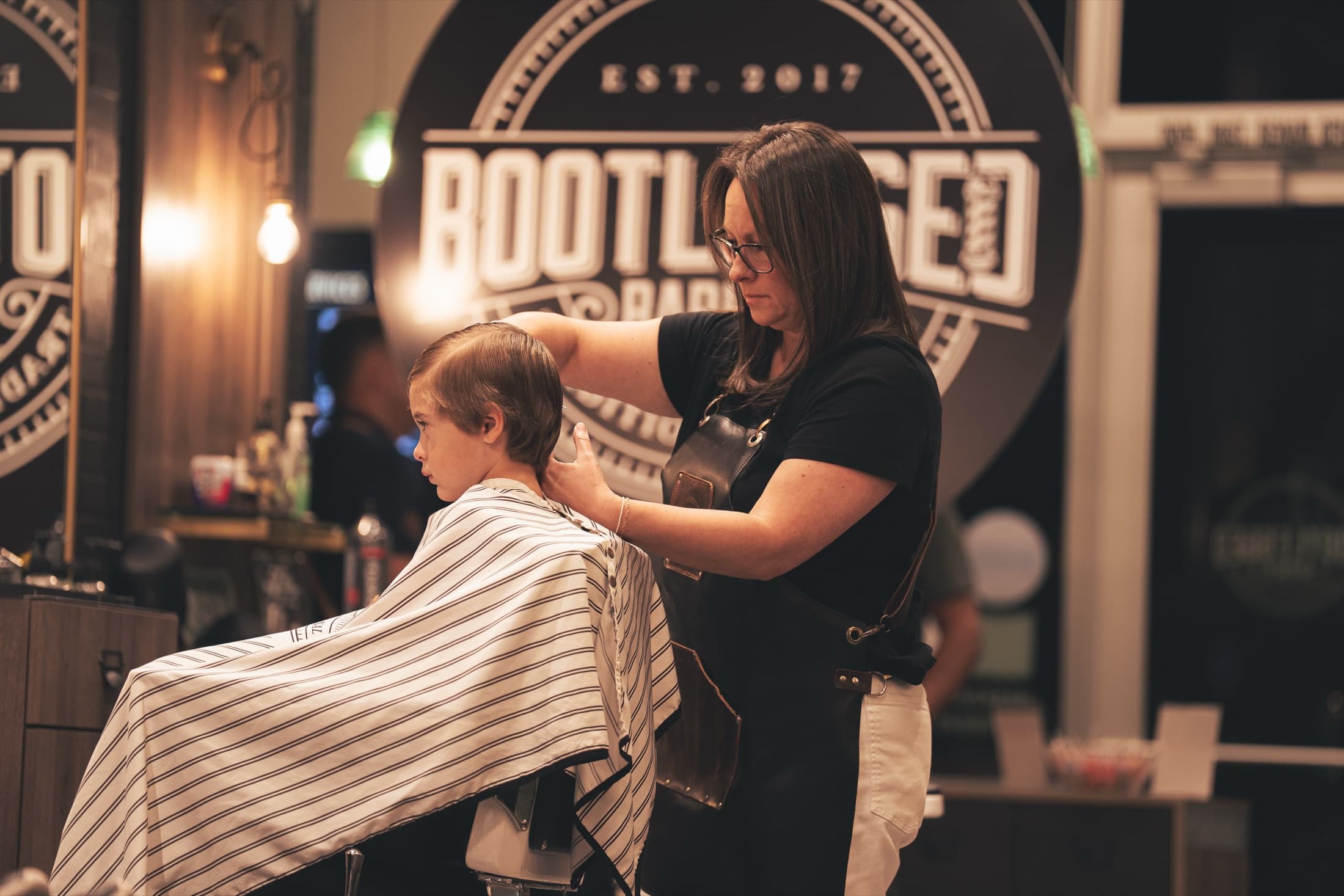 Barber cutting hair in front of the round Bootlegged Barber Est. 2017 wall sign in Herriman