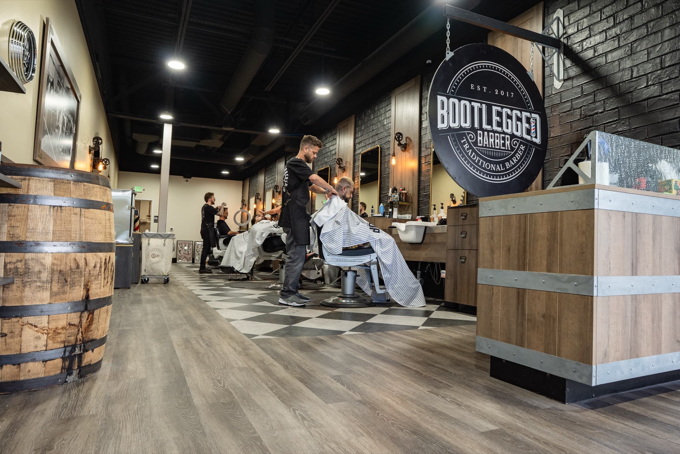 The Bootlegged Barber shop floor — whiskey barrel, round Est. 2017 sign, and a barber caping a client on the checkerboard floor